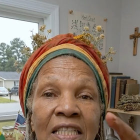 Photoreal close-up of Tia Ayanna, an older woman in a rastafari-color headwrap with dried flowers, speaking to camera